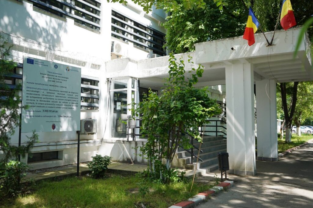spitalul caracal - Jurnalul Olteniei – Știri din Craiova și Oltenia White modern building entrance with a stairway, greenery, and two flags (Romania and another tricolor) on the roof; a large informational sign is posted to the left.