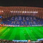 Night-time soccer match in a large stadium with fans forming blue and white cards in the stands and a fiery red banner display along one side of the field behind the goal, creating a dramatic atmosphere behind the pitch.