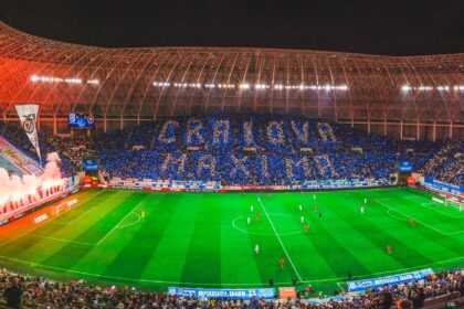 Night-time soccer match in a large stadium with fans forming blue and white cards in the stands and a fiery red banner display along one side of the field behind the goal, creating a dramatic atmosphere behind the pitch.