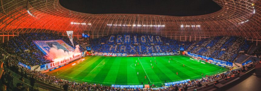 Night-time soccer match in a large stadium with fans forming blue and white cards in the stands and a fiery red banner display along one side of the field behind the goal, creating a dramatic atmosphere behind the pitch.