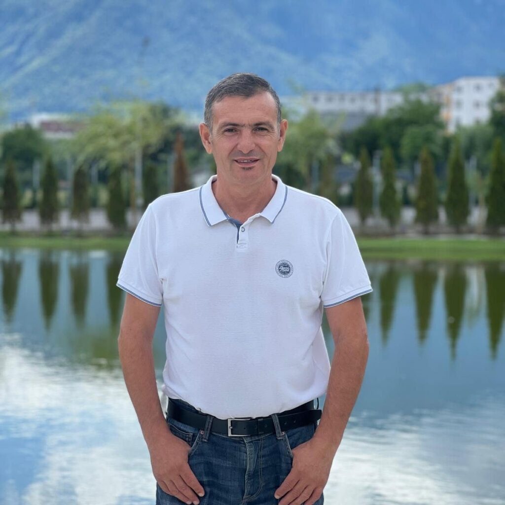 stefan popescu 518279339_2794033037448451_5389649669103612458_n - Jurnalul Olteniei – Știri din Craiova și Oltenia Smiling man in a white polo shirt stands by a lake, with trees and blue mountains in the background.