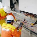 Two workers in high-visibility safety gear inspect solar inverter equipment and wiring on a rooftop, using a handheld tester.