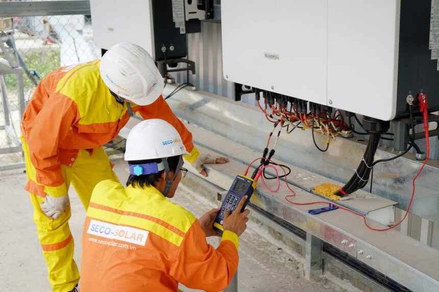 Two workers in high-visibility safety gear inspect solar inverter equipment and wiring on a rooftop, using a handheld tester.