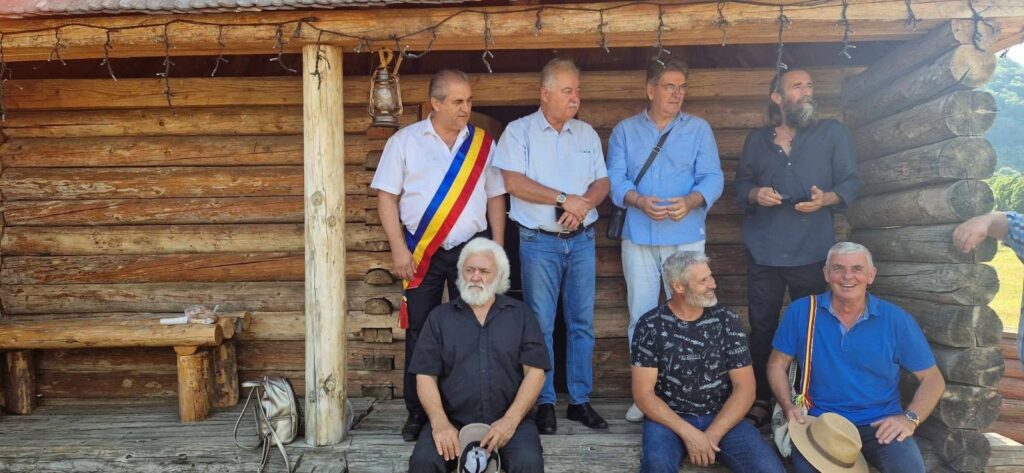 Group of seven men posing on a rustic log-cabin porch; one man wears a Romanian flag sash.