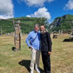 Two men stand together with arms around each other in a sunny outdoor park, wooden sculptures nearby and green hills in the background under a blue sky.