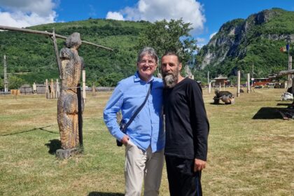 Two men stand together with arms around each other in a sunny outdoor park, wooden sculptures nearby and green hills in the background under a blue sky.