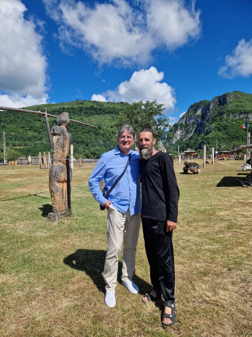 Two men stand together with arms around each other in a sunny outdoor park, wooden sculptures nearby and green hills in the background under a blue sky.