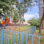 Playground in a residential yard with orange swings, colorful play structures, and a blue fence, under trees by a building with red-framed windows on the right.