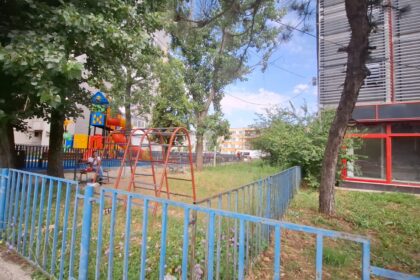 Playground in a residential yard with orange swings, colorful play structures, and a blue fence, under trees by a building with red-framed windows on the right.
