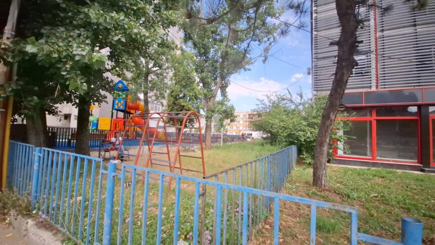Playground in a residential yard with orange swings, colorful play structures, and a blue fence, under trees by a building with red-framed windows on the right.