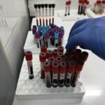 Gloved hand selecting a red-capped blood tube from a rack of specimen vials in a sterile lab.