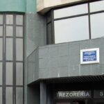 Facade of a gray stone building with large curved glass window on the left and a blue sign above the entrance that reads 'TREZORERIA SLATINA'.