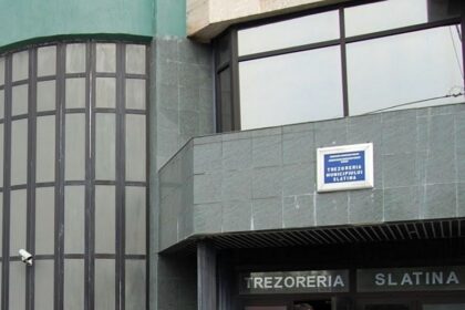 Facade of a gray stone building with large curved glass window on the left and a blue sign above the entrance that reads 'TREZORERIA SLATINA'.