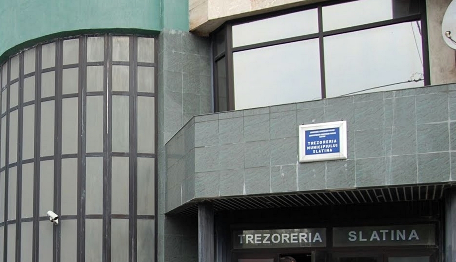 Facade of a gray stone building with large curved glass window on the left and a blue sign above the entrance that reads 'TREZORERIA SLATINA'.