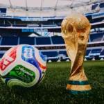 Soccer ball beside a golden FIFA World Cup trophy on a green stadium field with empty blue seats in the background (SoFi Stadium).