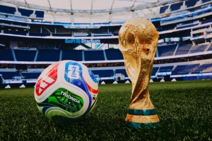 Soccer ball beside a golden FIFA World Cup trophy on a green stadium field with empty blue seats in the background (SoFi Stadium).