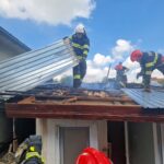 Firefighters on a damaged roof lifting a corrugated metal panel amid light smoke and a blue sky above.