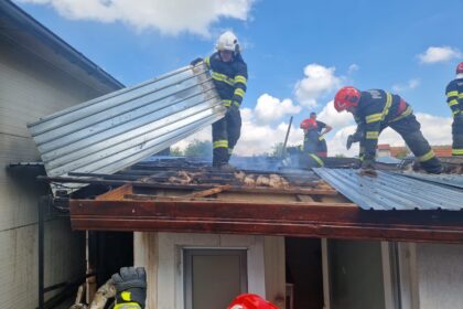 Firefighters on a damaged roof lifting a corrugated metal panel amid light smoke and a blue sky above.