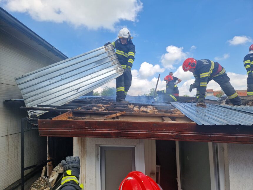 Firefighters on a damaged roof lifting a corrugated metal panel amid light smoke and a blue sky above.
