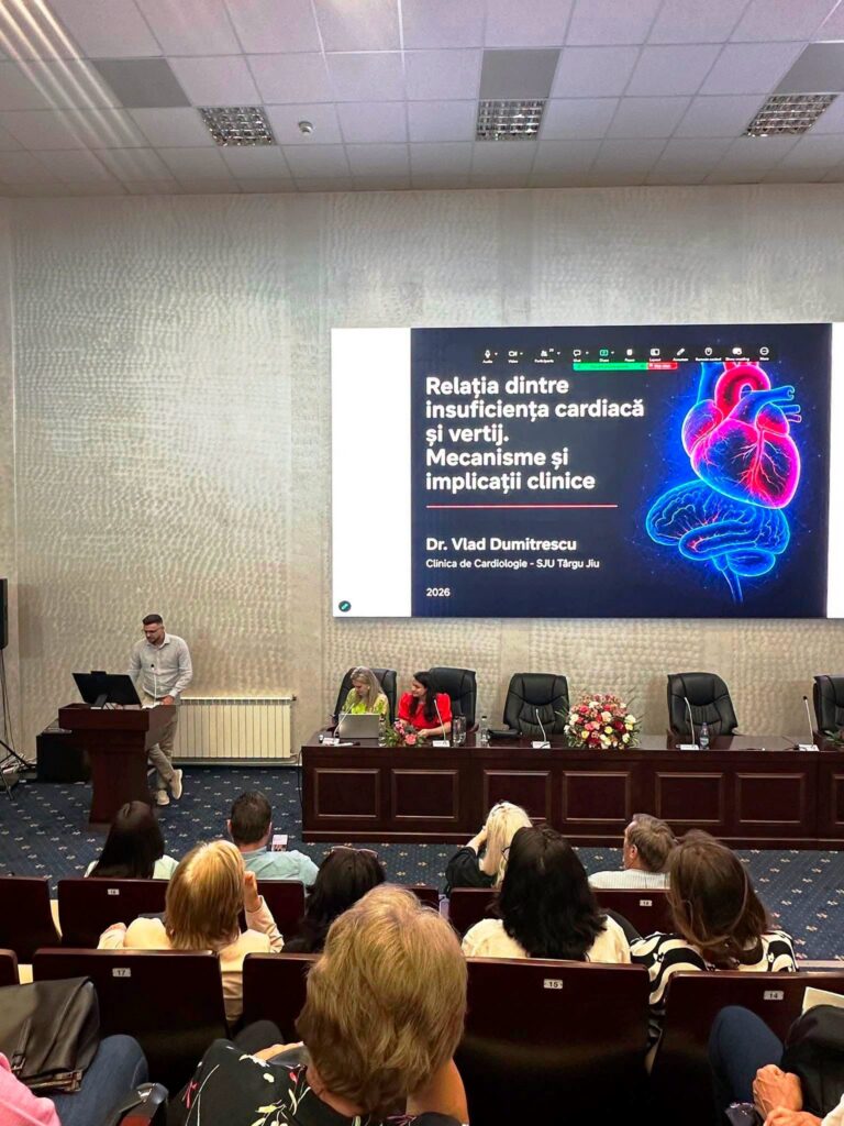 vertij proiect - Jurnalul Olteniei – Știri din Craiova și Oltenia Speaker at a podium presenting to an audience in a conference room, with a large poster about heart health on the wall behind.