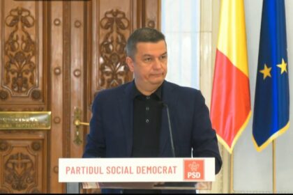 Man in a dark blazer speaks at a podium labeled 'Partidul Social Democrat' with Romanian and EU flags in the background.