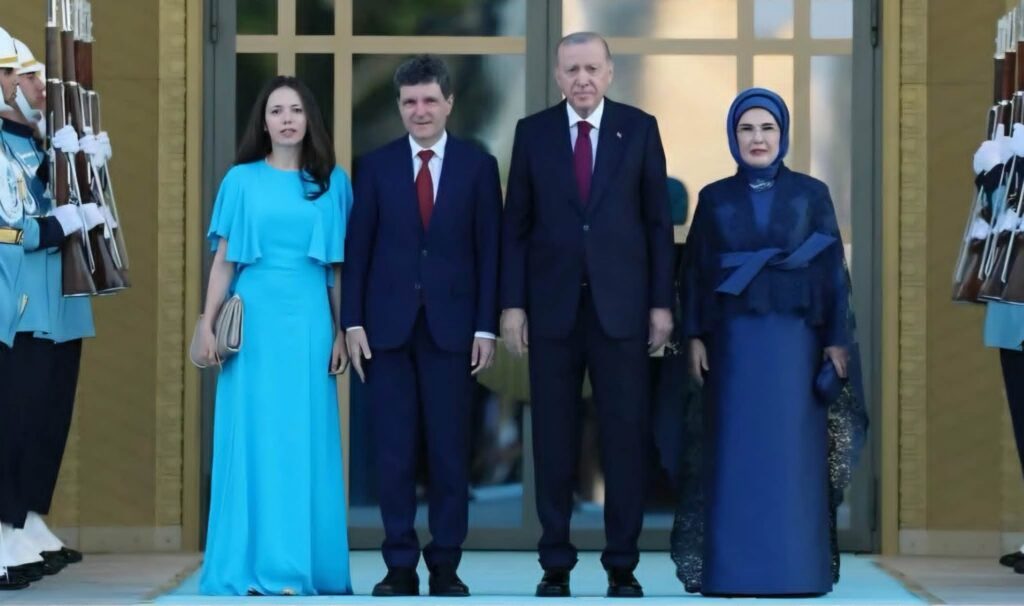 Four formally dressed people pose at a doorway, two men in suits and two women in blue outfits, flanked by guards on both sides of the entrance.