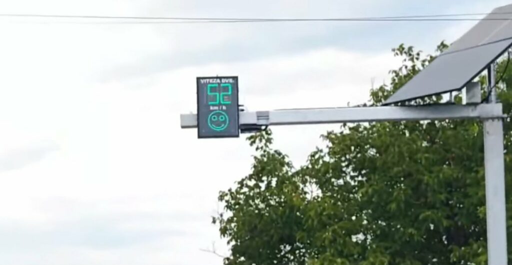 Electronic speed display sign showing '52' with a green smiley face on a metal arm near a tree and solar panel in the background.