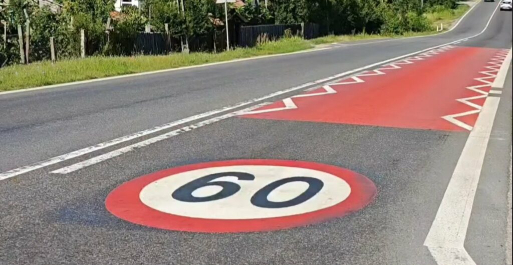 Large red speed zone painted on the road with a white 60 circle, adjacent to a long red area outlined by white chevrons along the curb.
