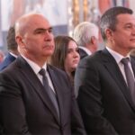 Two men in dark suits stand at a formal ceremony, facing forward, with other attendees blurred in the background.