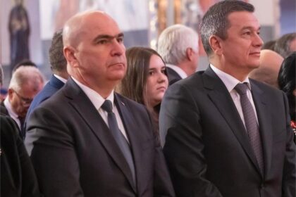 Two men in dark suits stand at a formal ceremony, facing forward, with other attendees blurred in the background.