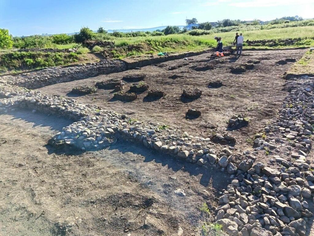 Archaeological dig site with multiple circular stone-lined pits in a dirt field; a low stone wall encircles the area, and three people are in the background.