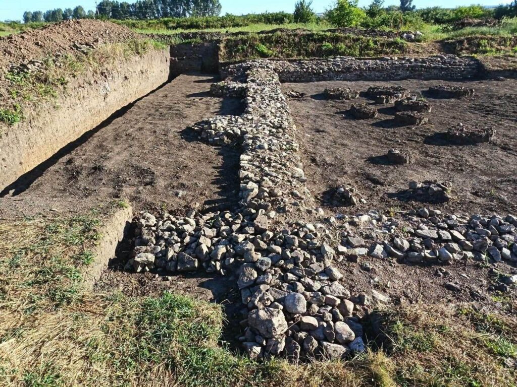Archaeological site with low stone walls and circular sunken pits in bare earth, surrounded by grass and shrubs.