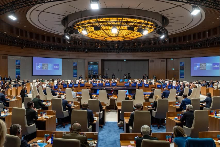 Round conference hall filled with delegates around a central dais for a NATO meeting in progress