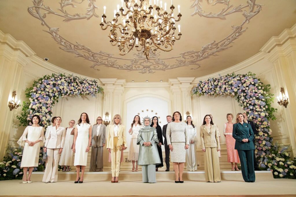Group of women in elegant pastel and neutral outfits standing in a grand, decorated room with a large chandelier and floral arches behind them.