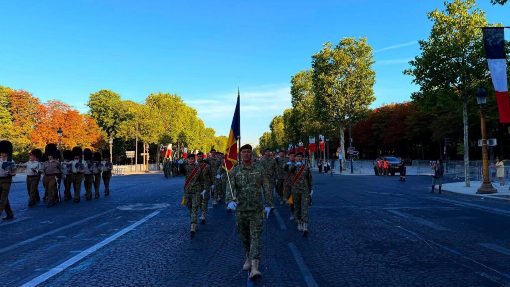 - Jurnalul Olteniei – Știri din Craiova și Oltenia Soldiers march in formation down a cobblestone street during a parade, led by a flag bearer with a blue-yellow-red flag, autumn trees lining the route.