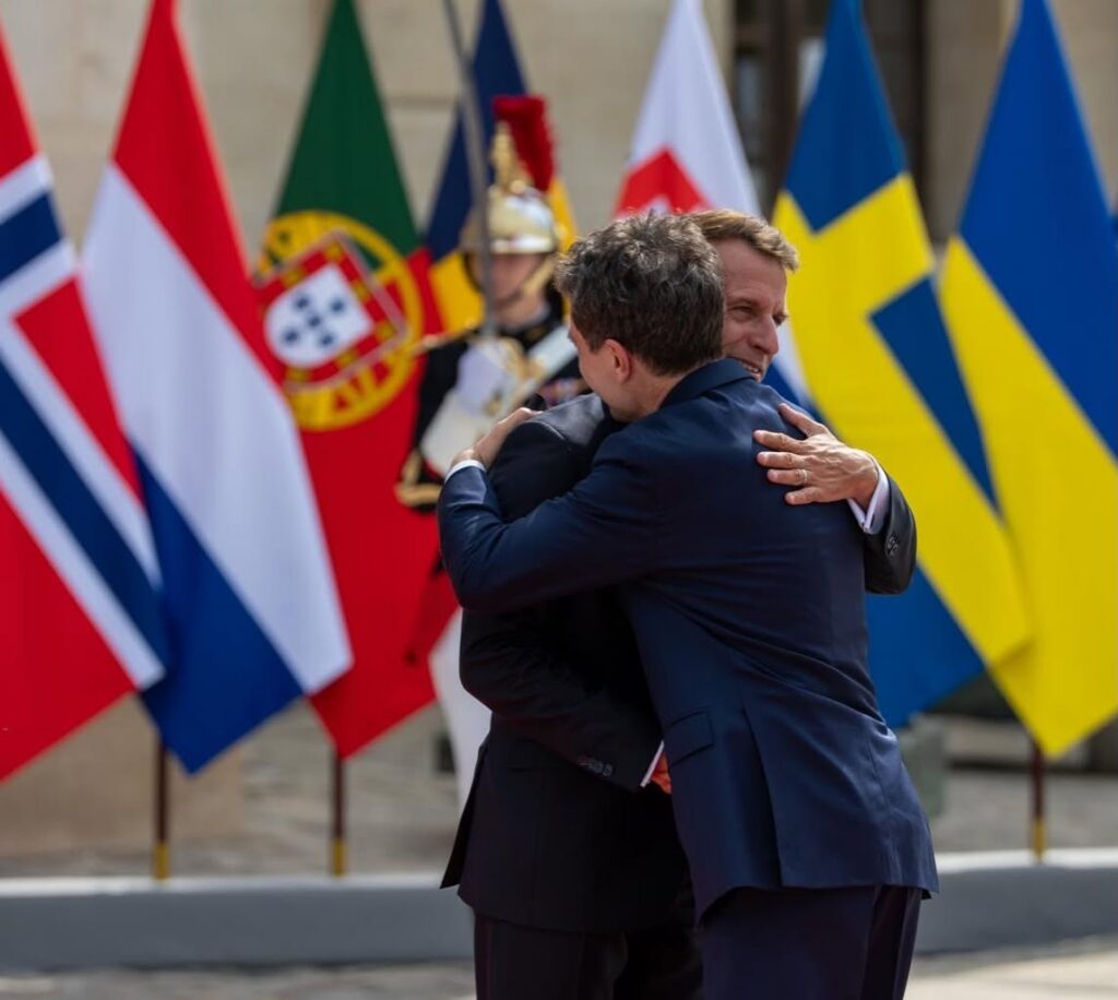 Two men in dark suits embrace in a formal moment with international flags in the background.