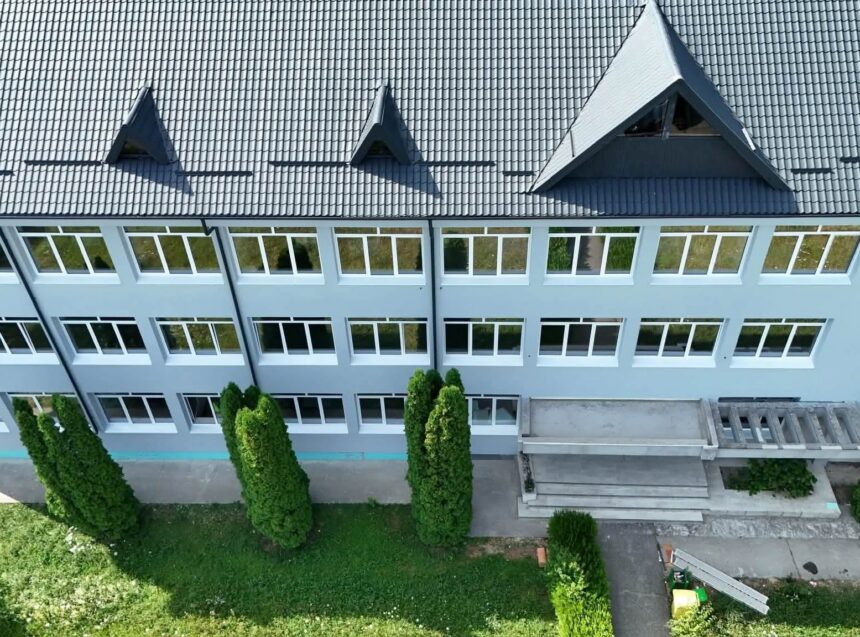 Aerial view of a pale blue, multi-story building with many white-framed windows, gray tiled roof, and neatly trimmed evergreens below.