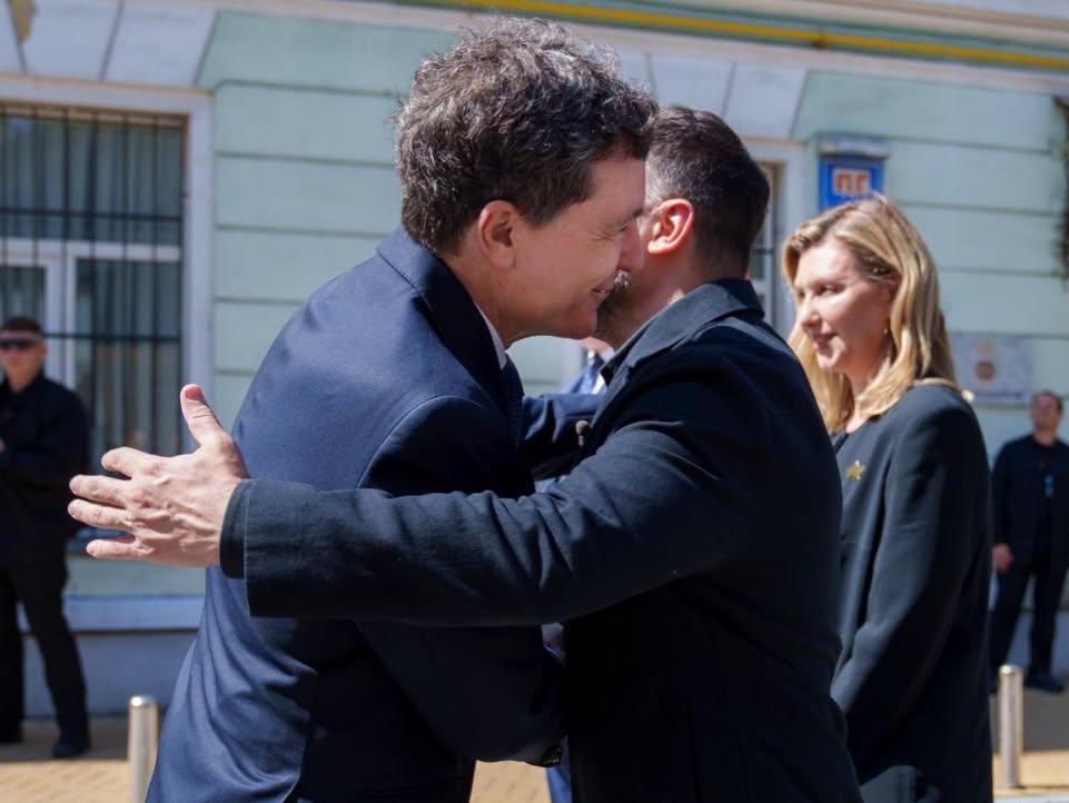 Two men in suits embrace in a friendly hug outdoors while a woman watches nearby.