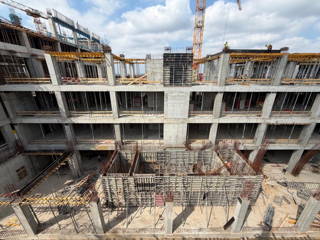 Under-construction concrete building with scaffolding, steel rebar, and workers on the top floor; a crane rises in the background.