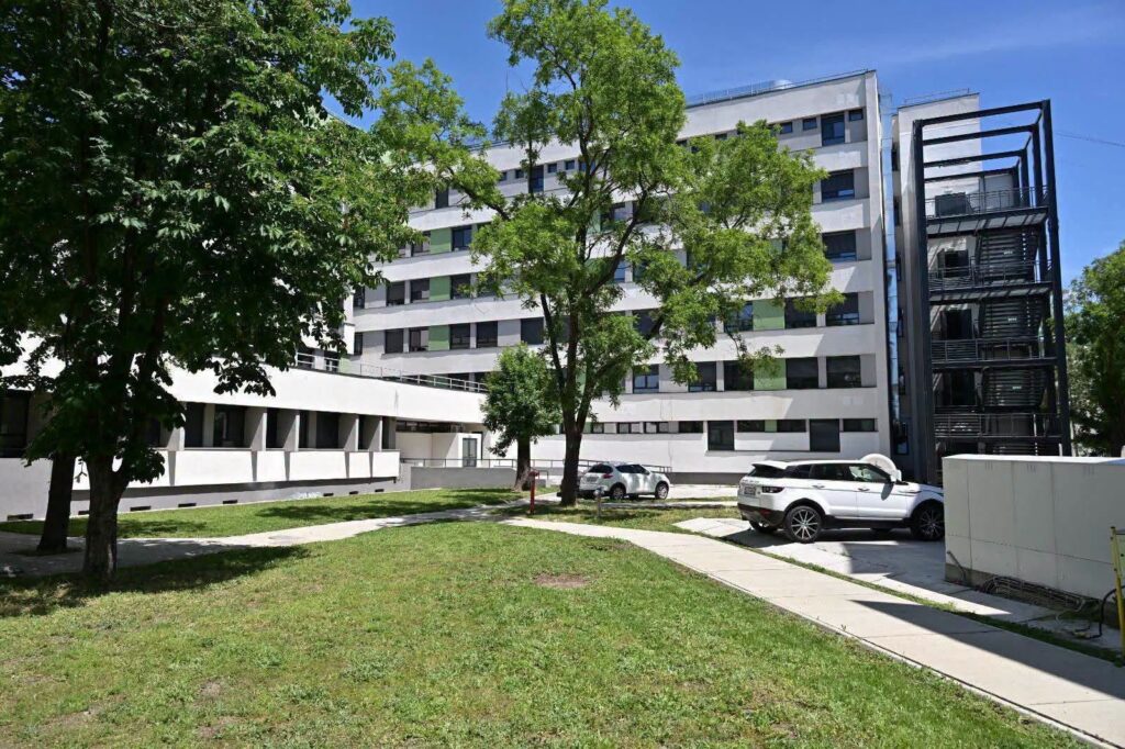 White multi‑story residential building with green window shades, set in a tree-filled courtyard and grassy lawn.