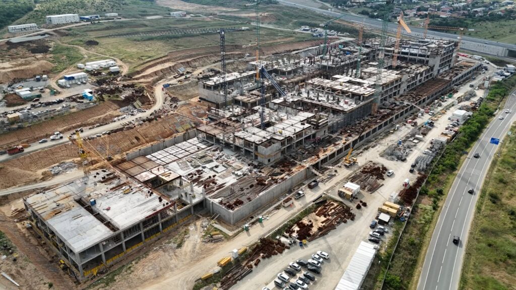 Aerial view of a large construction site with cranes and partially built concrete structures beside a highway on the right edge.