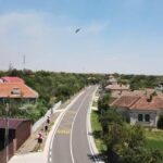 Curved rural street through a small village with red-tiled roofs, greenery, and a bird flying in a clear blue sky above.