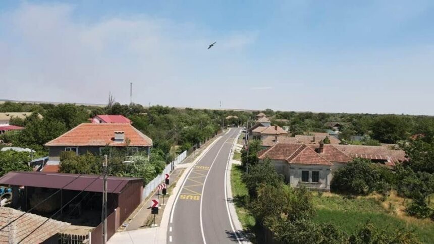 Curved rural street through a small village with red-tiled roofs, greenery, and a bird flying in a clear blue sky above.