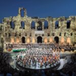 Evening concert inside a historic stone amphitheater, with a large choir and orchestra on a circular stage surrounded by stone arches and audience.