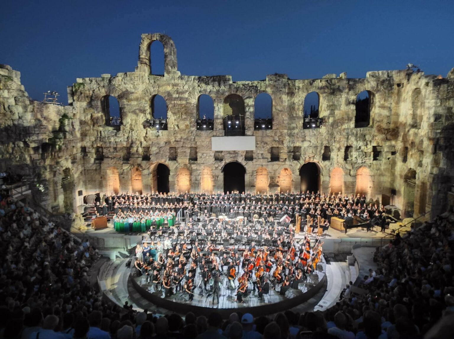 Evening concert inside a historic stone amphitheater, with a large choir and orchestra on a circular stage surrounded by stone arches and audience.