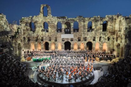Evening concert inside a historic stone amphitheater, with a large choir and orchestra on a circular stage surrounded by stone arches and audience.