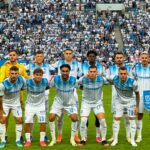 Soccer team in white-and-blue striped kits posing for a pre-match group photo on the field with a packed stadium behind them