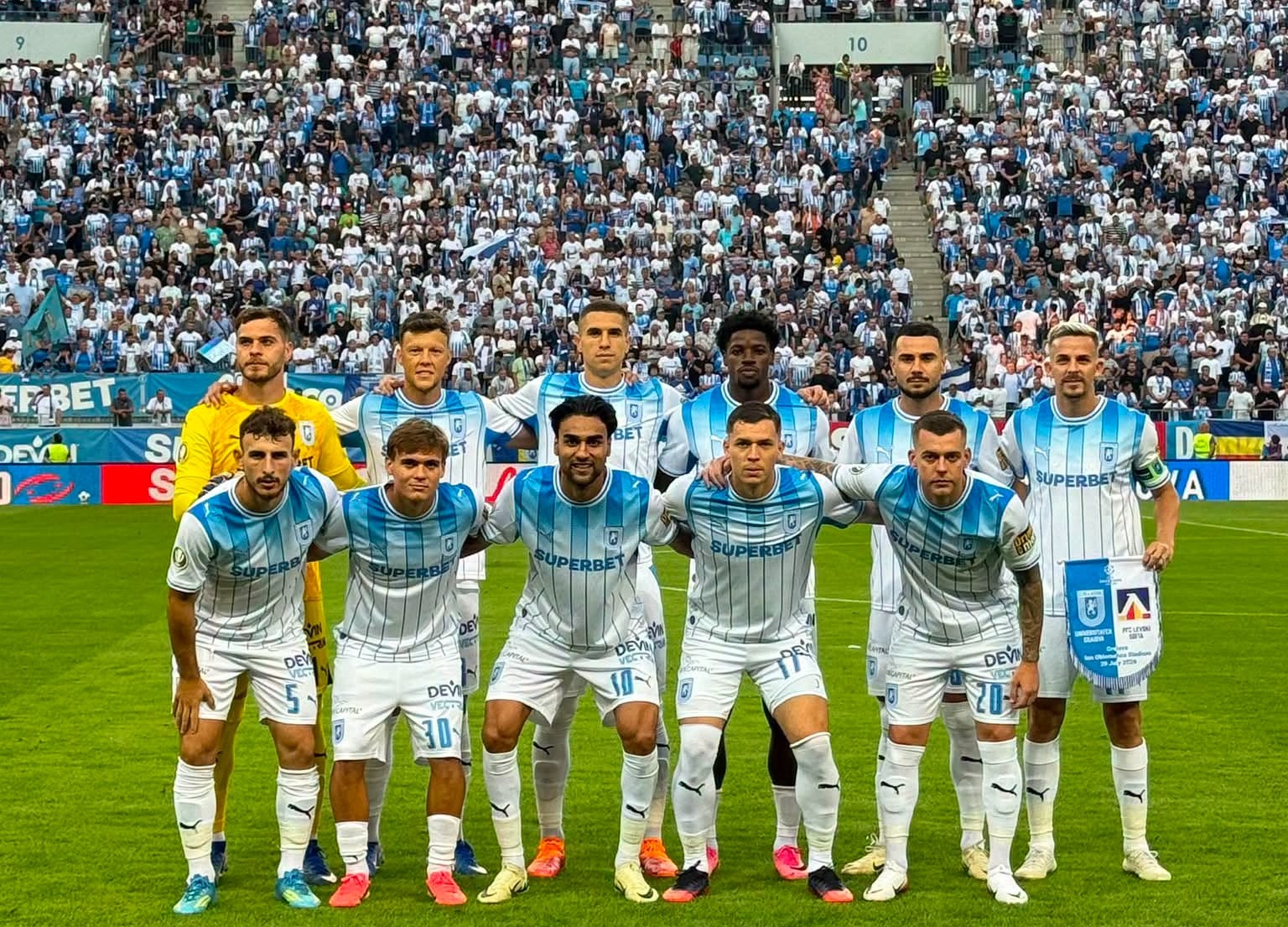 Soccer team in white-and-blue striped kits posing for a pre-match group photo on the field with a packed stadium behind them