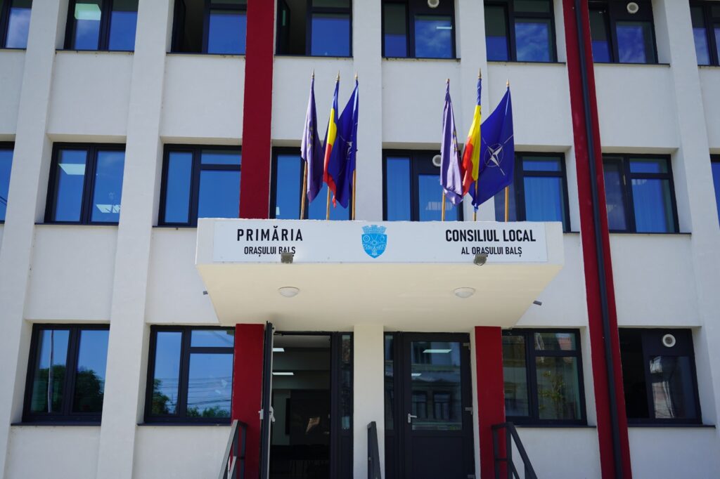 Front of Balș City Hall with a row of national/European flags and a sign reading 'Primăria Orașului Balș' and 'Consiliul Local' with a blue crest above the entrance