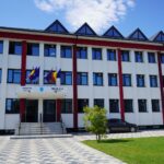 City hall building with white facade and red columns, flags at the entrance and a sign for Primăria/Consiliul Local under a blue sky.